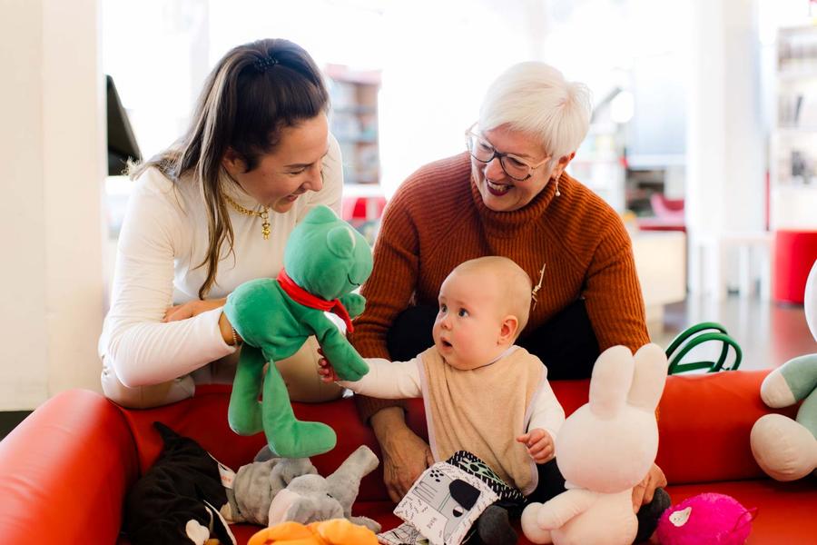 Een baby zit tussen knuffels op een rode bank en reikt naar een groene Kikker handpop, terwijl twee volwassenen ernaast zitten en samen interactie hebben tijdens een babybiebbijeenkomst.
