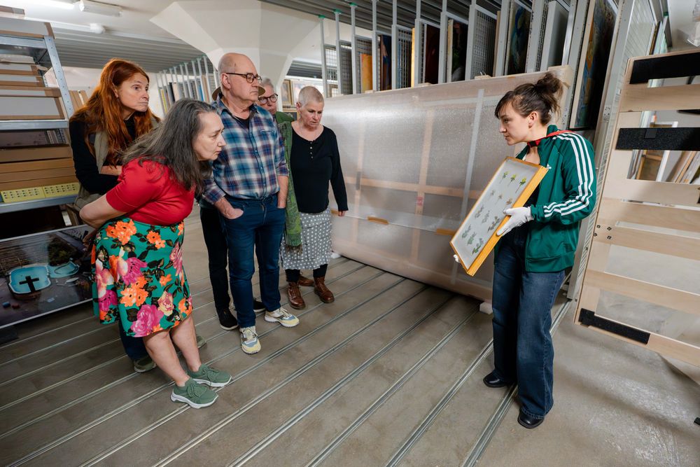 Groep mensen luistert naar uitleg van een medewerker die een ingelijst werk toont in het depot.