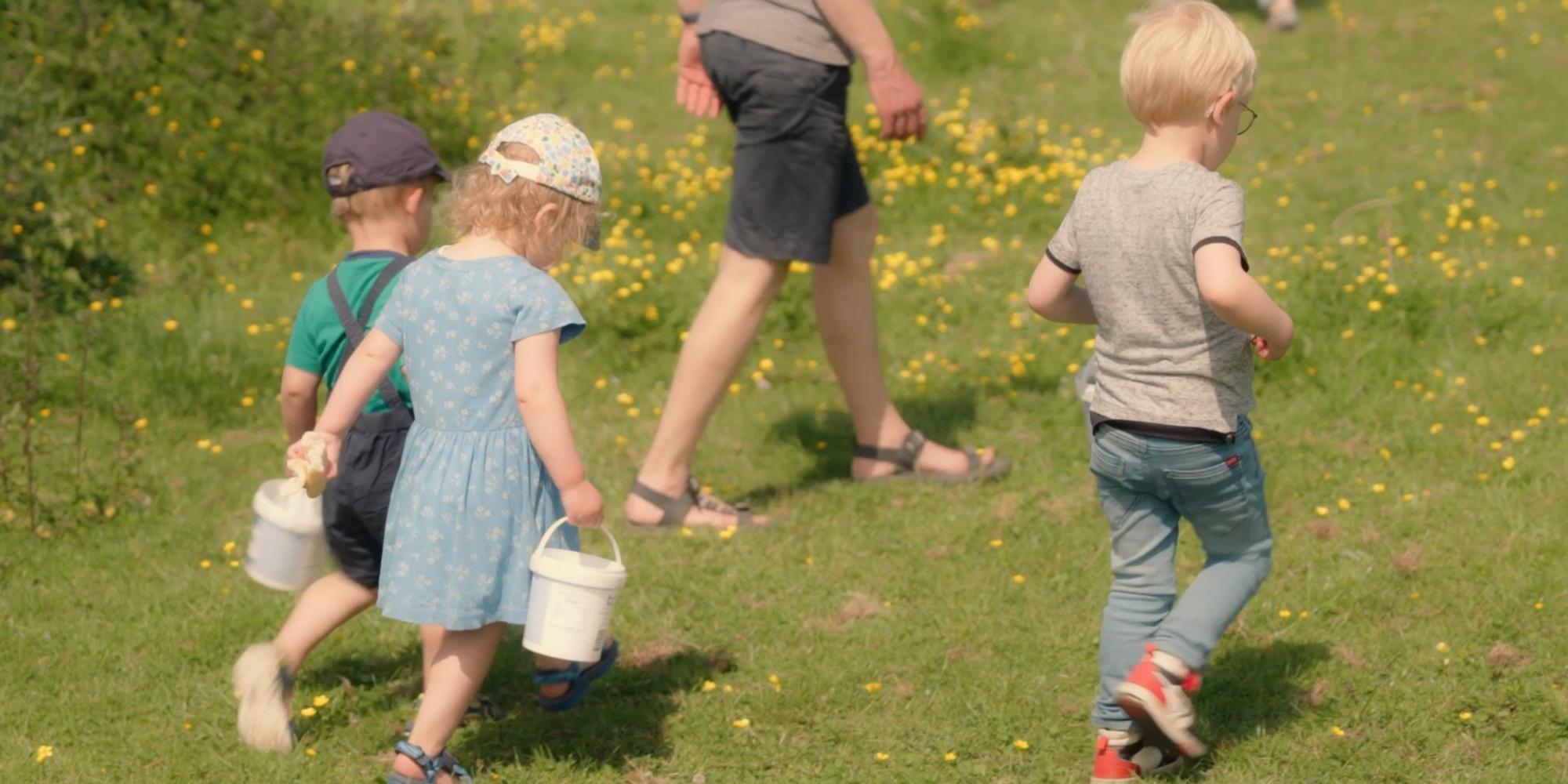 Jonge kinderen lopen over een grasveld met gele bloemen, sommigen dragen kleine witte emmertjes. Een volwassene loopt voor hen uit.