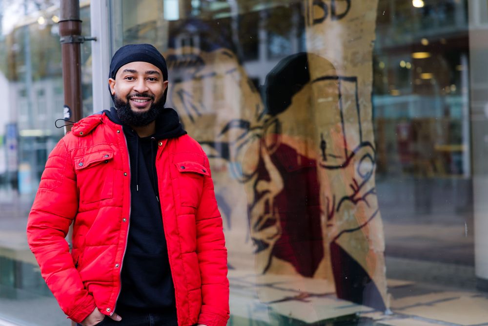 Kunstenaar Quentley Barbara poseert lachend in een rode jas voor zijn kartonnen kunstwerk in de vitrine van SCHUNCK in Heerlen.