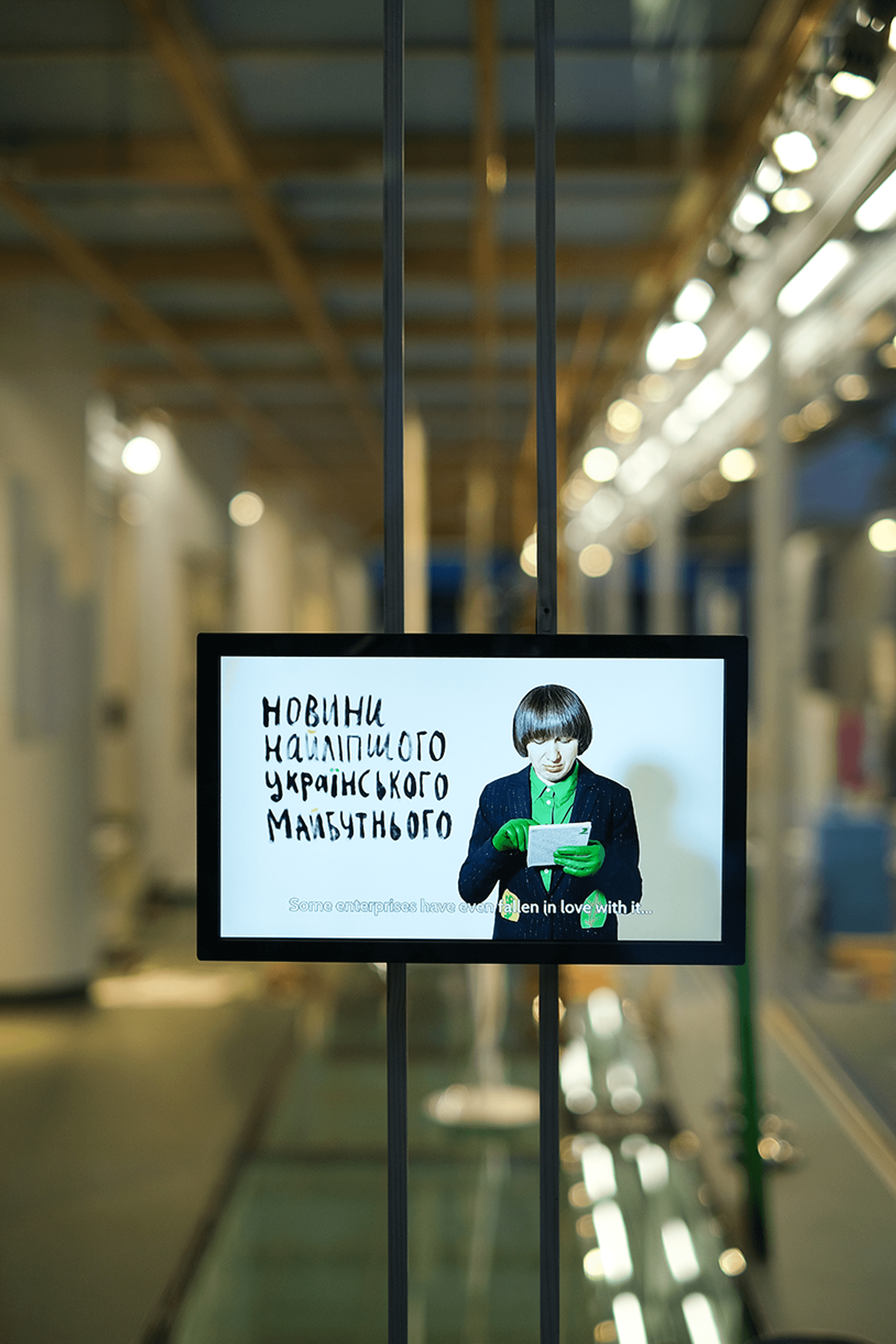 Een beeldscherm toont een vrouw met kort bruin haar, een groene blouse en groene handschoenen, die een papier leest; links naast haar staat Oekraïense tekst geschreven.