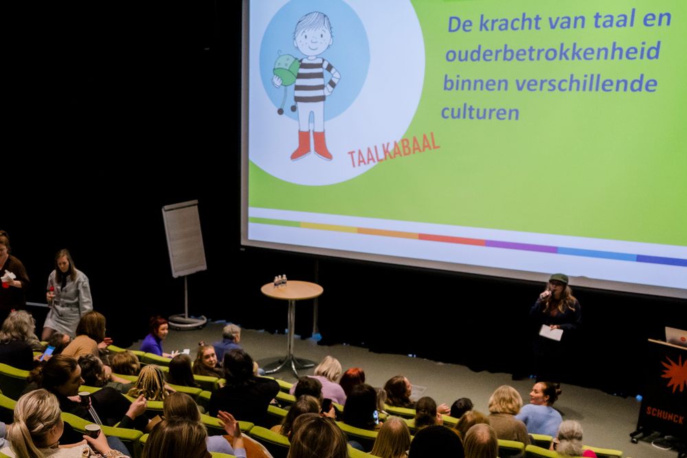 Een vol auditorium in SCHUNCK met mensen in groene stoelen kijkt naar een presentatie op een groot scherm met de titel “De kracht van taal en ouderbetrokkenheid binnen verschillende culturen”, onderdeel van het programma “Taalkabaal”.
