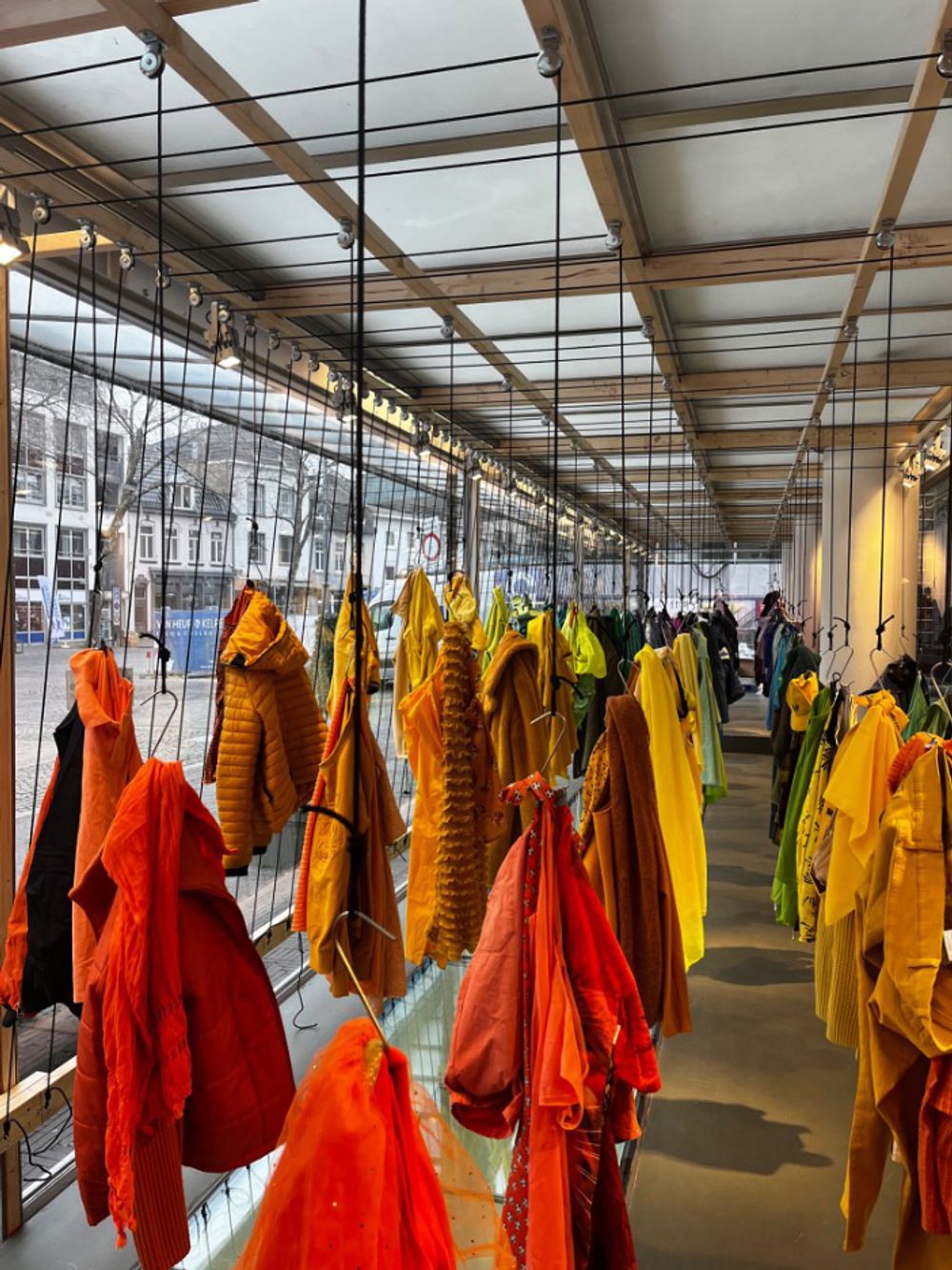 Binnenzicht in de vitrine van SCHUNCK, waar felgekleurde kledingstukken in rood, oranje, geel en groen aan haken hangen in een strakke opstelling, onderdeel van een kunstinstallatie.