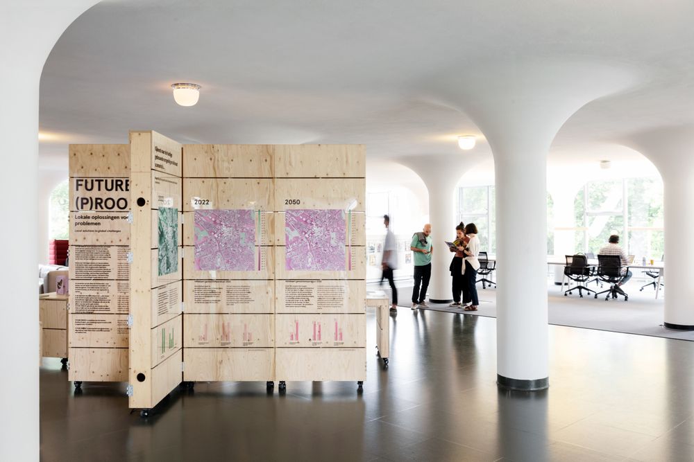 Tentoonstelling Future (P)roof in SCHUNCK met houten panelen waarop kaarten van 2022 en 2050 zijn afgebeeld, met uitleg. Op de achtergrond bekijken bezoekers samen de informatie in de lichte, open ruimte van het Glaspaleis.