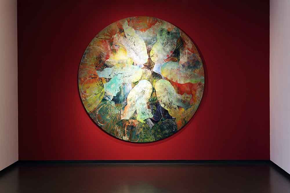 Rond schilderij met gestileerde, overlappende lichamen in pastel- en aardetinten tegen een rode muur. Het werk heeft een gelaagde, textuurrijke uitstraling en een dynamische compositie.