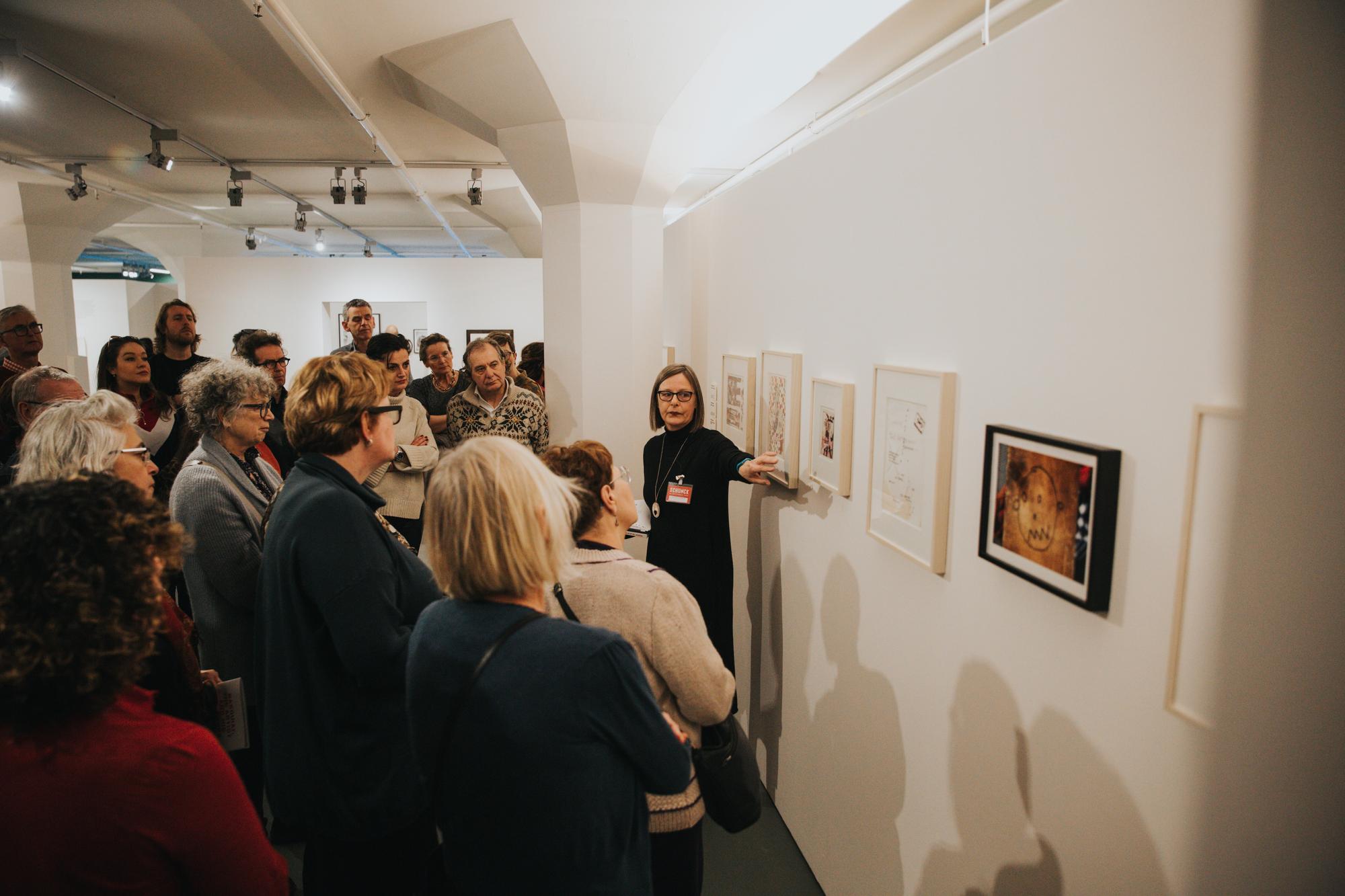 Rondleiding in een kunstgalerie waarbij een gids een groep bezoekers uitleg geeft over de kunstwerken aan de muur.