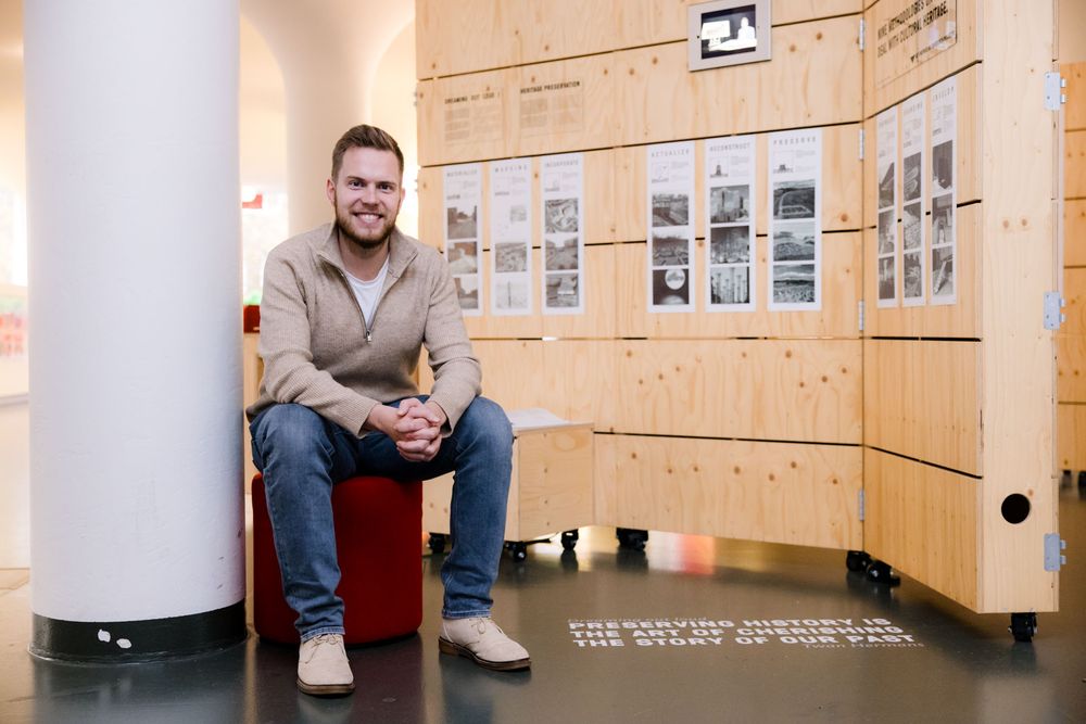 Portret Twan Hermans zittend op een kruk voor een houten presentatie-meubel waar foto's ophangen.