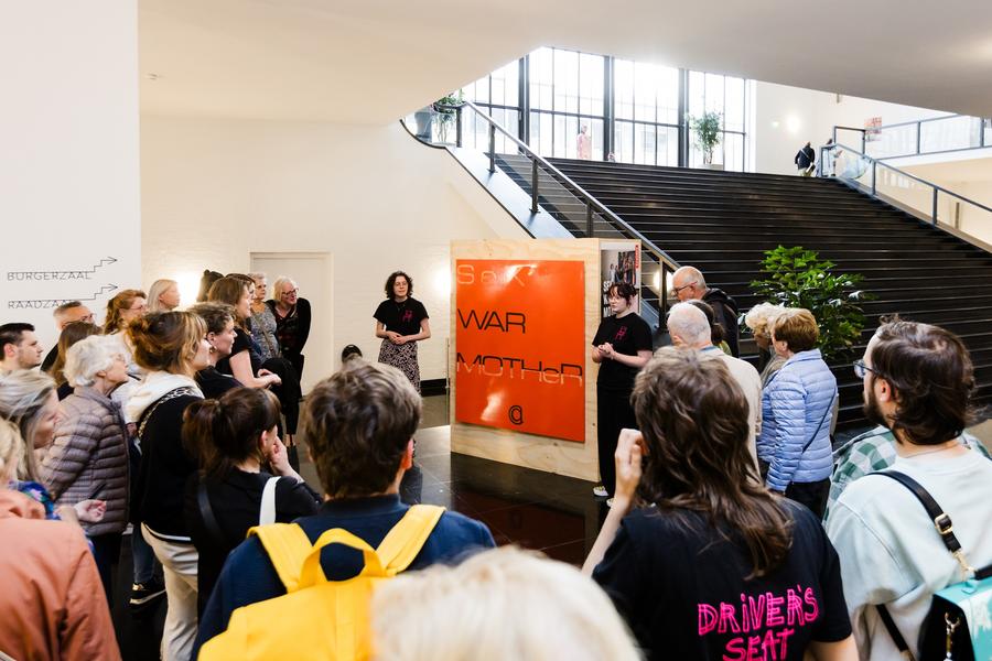 Twee leden van Driver's Seat staan bij een schilderij en geven een rondleiding aan een grote groep mensen. Op de achtergrond is een grote trap te zien. Het schilderij toont een oranje vlak met de tekst: "Sex, War, Mother".