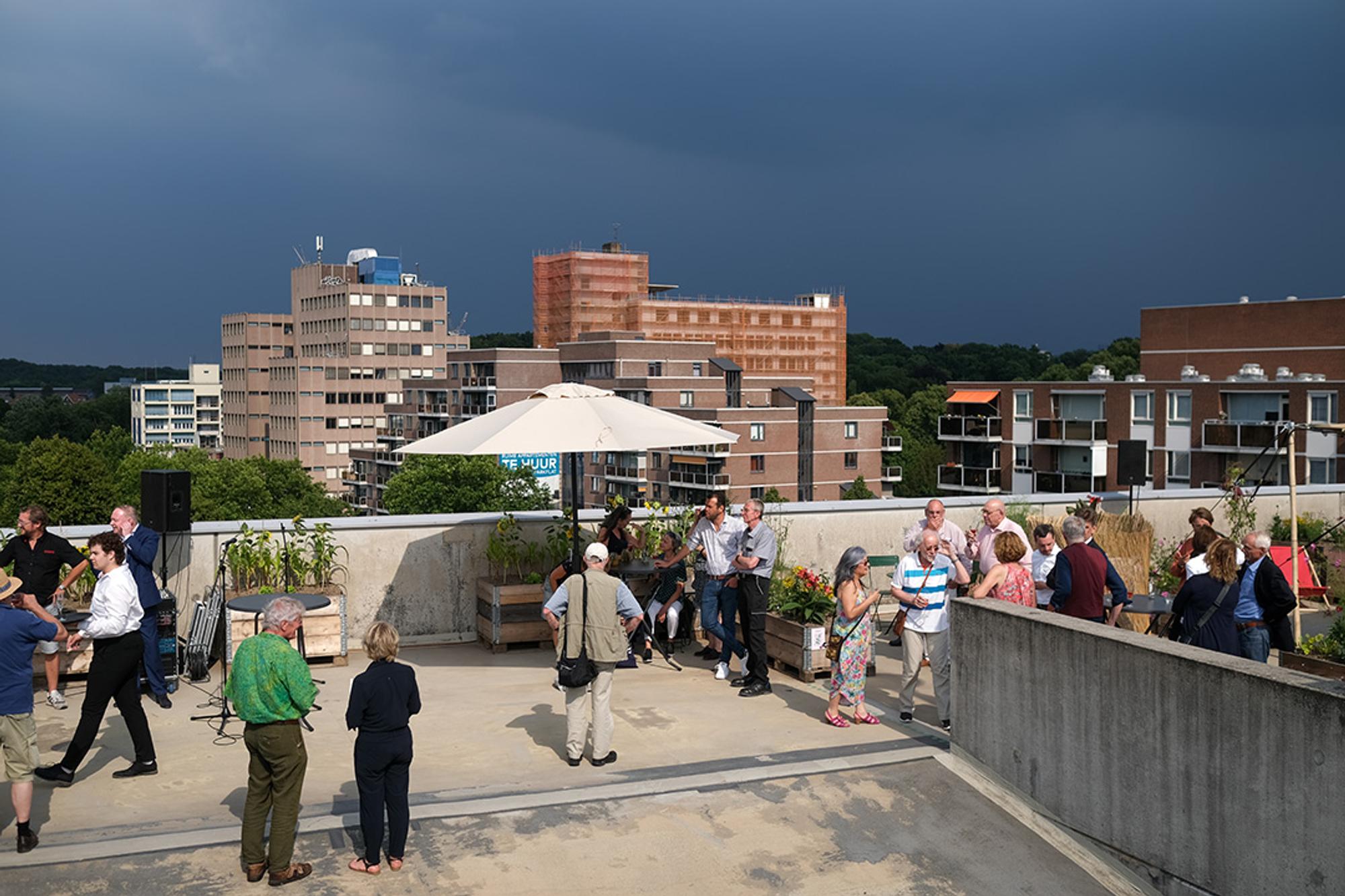 Een groep mensen verzamelt zich op een daktuin in een stedelijke omgeving, onder een grote witte parasol, omringd door houten plantenbakken met bloemen en groenten, met een dreigende donkere lucht op de achtergrond.