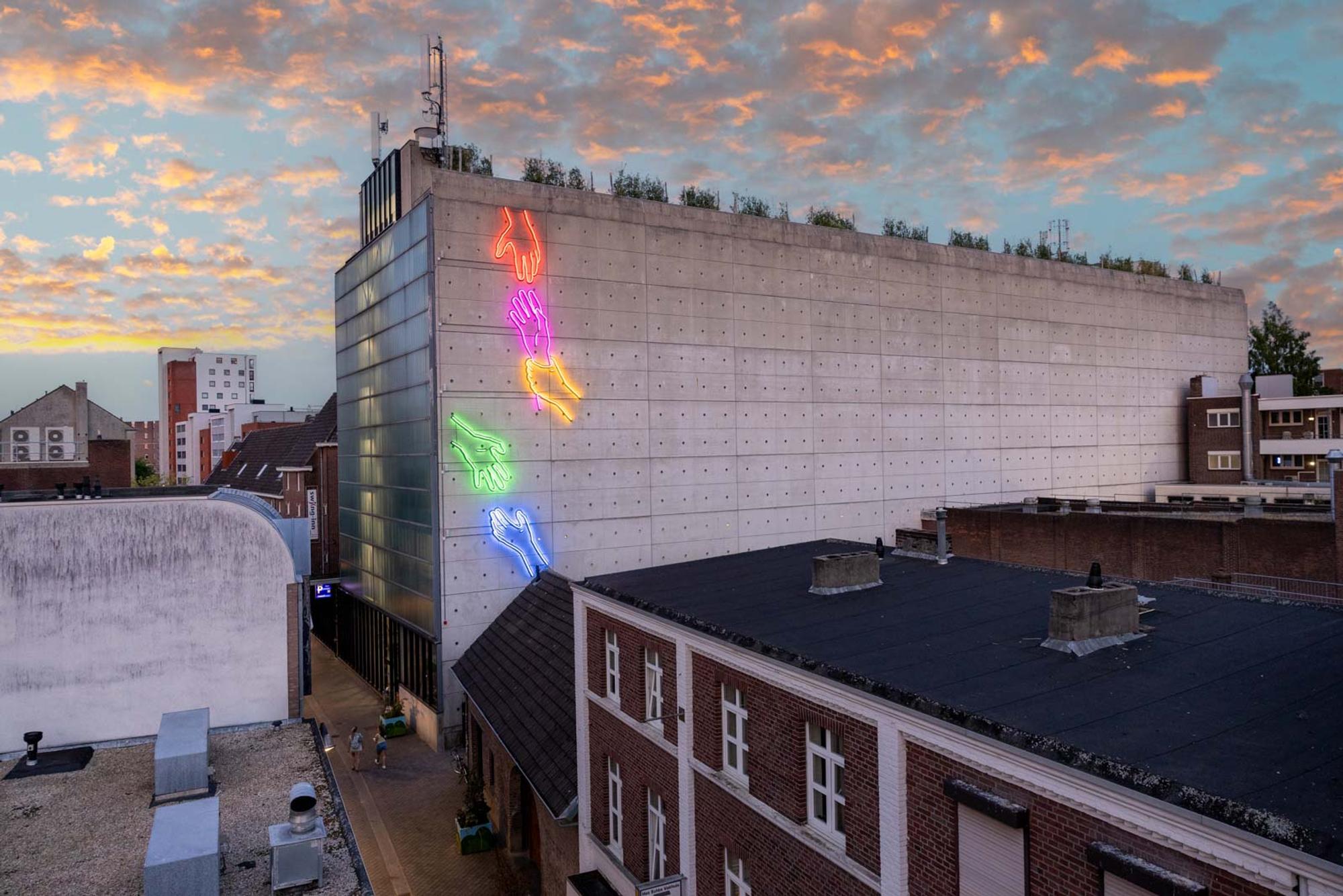 Stadsgezicht bij zonsondergang met een groot betonnen gebouw versierd met kleurrijke neoncontouren van dieren, waaronder een rode vogel, roze hand, gele vis, groene konijn en blauwe hond. De lucht is gevuld met wolken die worden verlicht door de ondergaande zon, omringd door gebouwen in diverse architectonische stijlen.