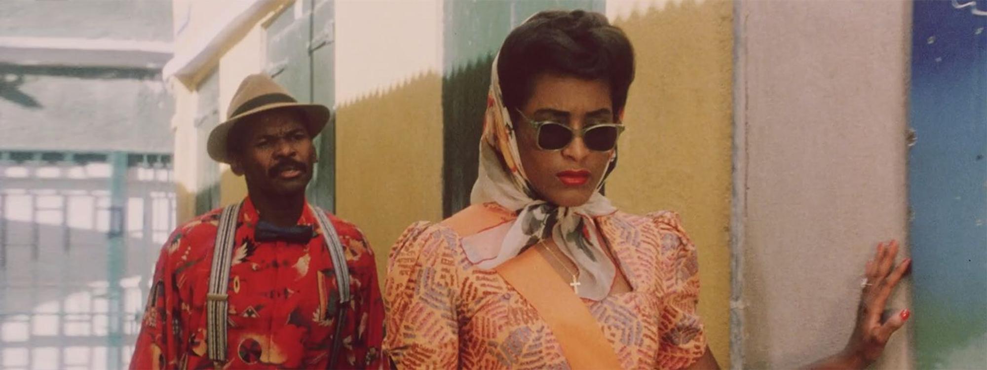 Een man en een vrouw wisselen een glimlach in een kleurrijke straat op Curaçao. Beiden dragen stijlvolle retrokleding en zonnebrillen. Still uit de film Ava & Gabriel (1990).