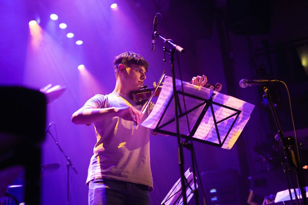 Op een podium tijdens een optreden van SCHUNCK Academy staat een muzikant voor een muziekstandaard en bespeelt een viool met een strijkstok. De scène is verlicht met paarse en roze podiumlichten, met microfoons en andere instrumenten zichtbaar op de achtergrond.
