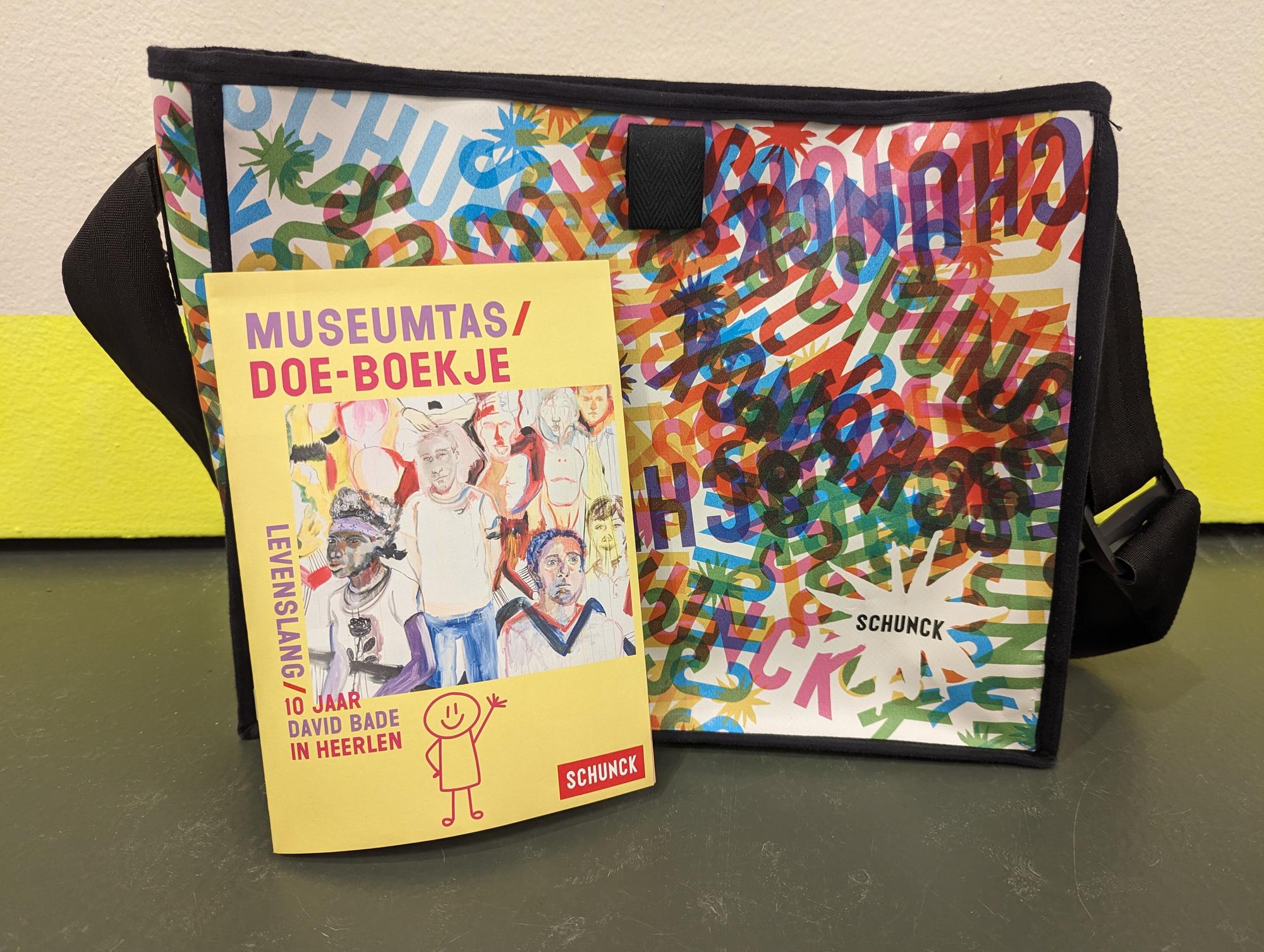 Museumtas met SCHUNCK-logo in kleurrijk patroon, met daarvoor het Museumtas doe-boekje waarop te lezen is: “Museumtas doe-boekje, Levenslang, 10 jaar David Bade in Heerlen.”
