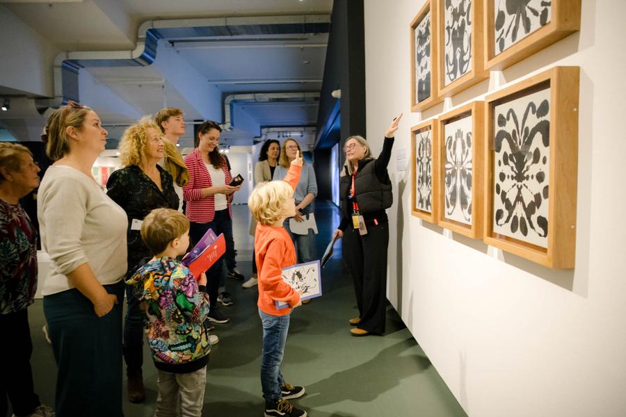 Een aantal ouders en hun kinderen staan in de museumzaal voor een aantal kunstwerken van Andy Warhol. De rondleidster vertelt hen een en ander over de werken waar zij naar kijken. 