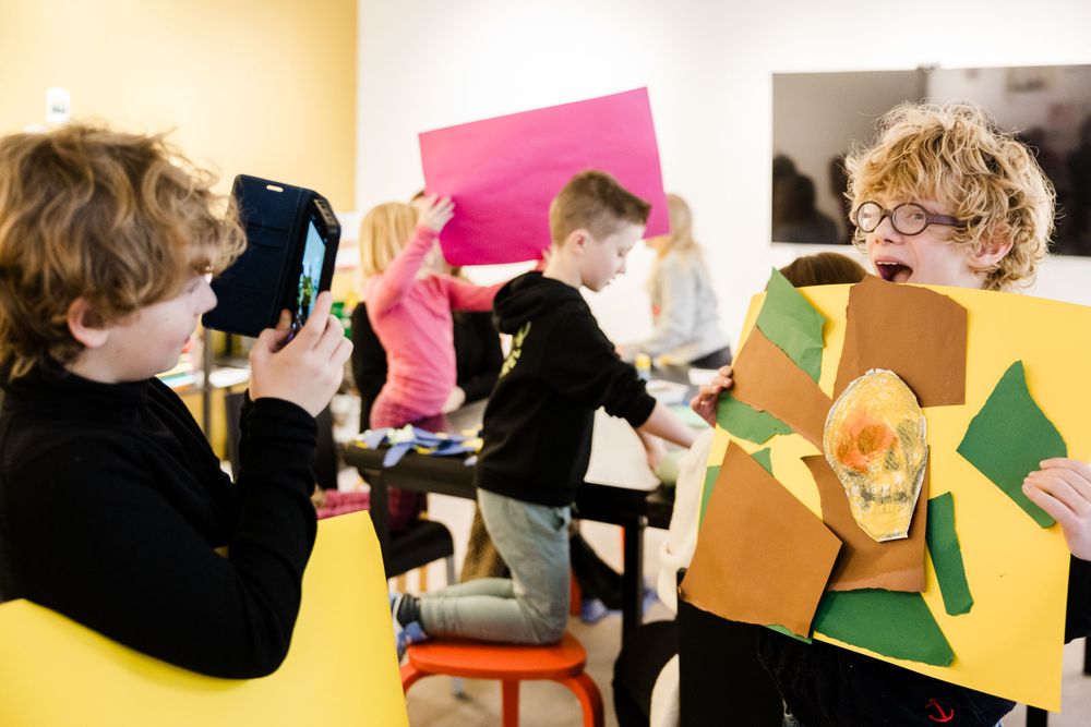 Kinderen werken creatief samen in een klaslokaal. Op de voorgrond fotografeert een kind een kleurrijk kunstwerk van karton en papier dat door een ander kind trots wordt gepresenteerd, terwijl op de achtergrond druk wordt geknutseld en geëxperimenteerd.