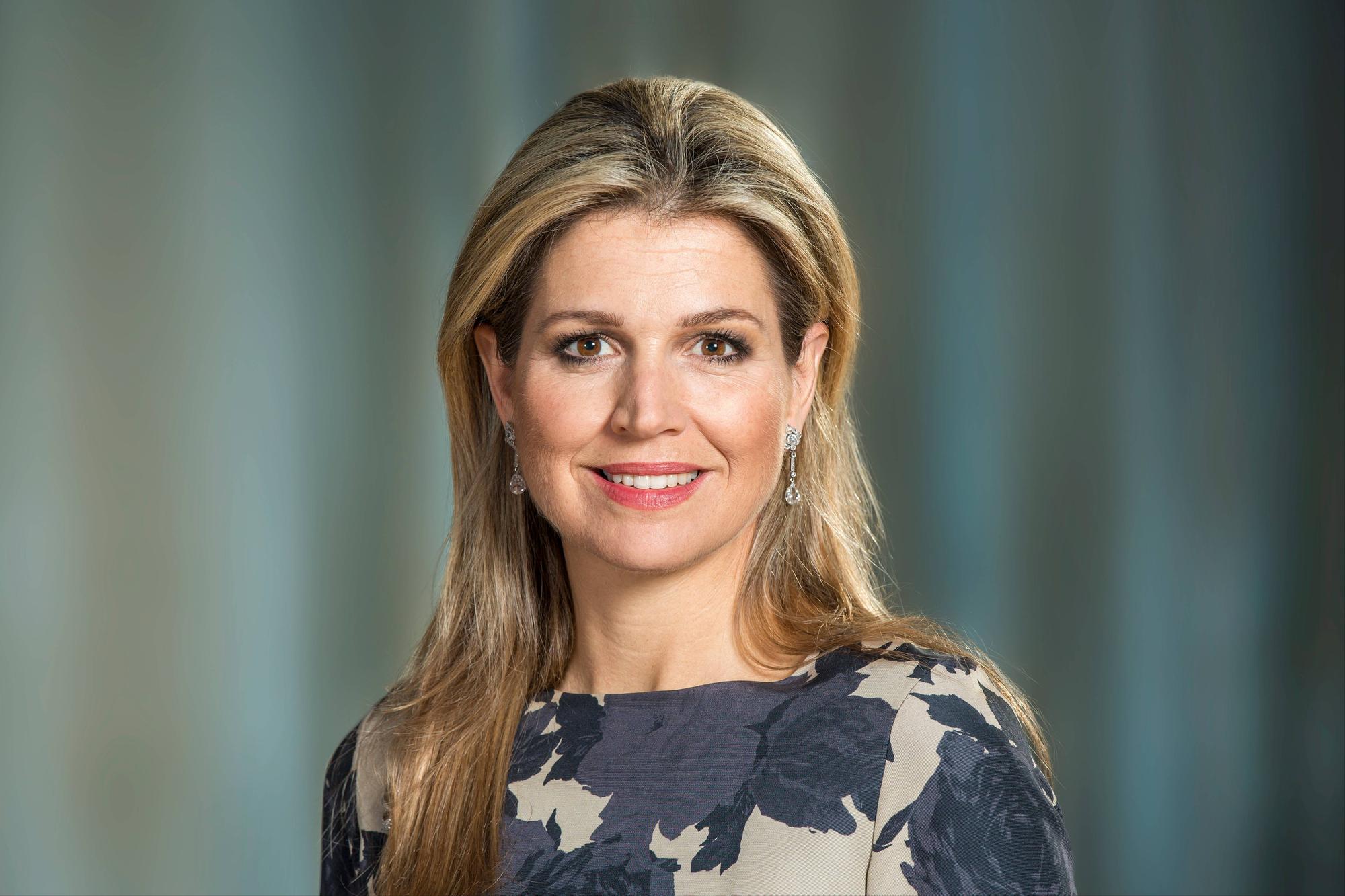 koningin Máxima met een glimlach, gekleed in een bloemrijke jurk met oorbellen, tegen een zachte, blauwe achtergrond.
