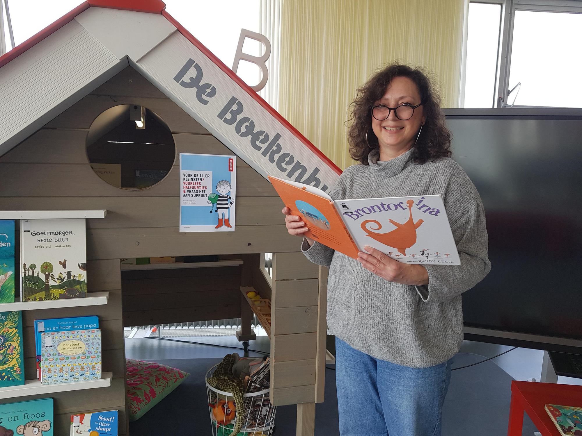 Portret vrijwilliger Annemiek Triepels Bibliotheek, met een boek in de handen staat ze voor een huisje met boeken genaamd "de boekenhut". 