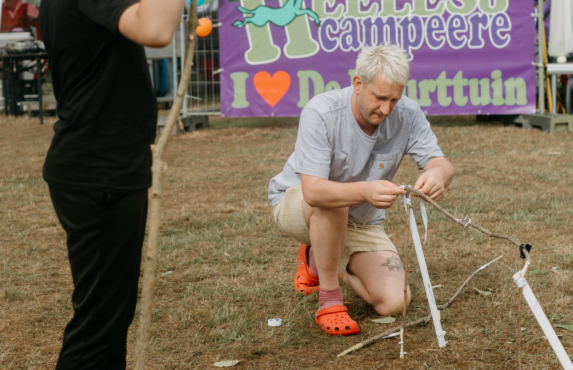 Kunstenaar Maarten Bel knielt op een grasveld en bevestigt witte linten aan takken tijdens een activiteit op de Buurtcamping in Heerlen-Noord.