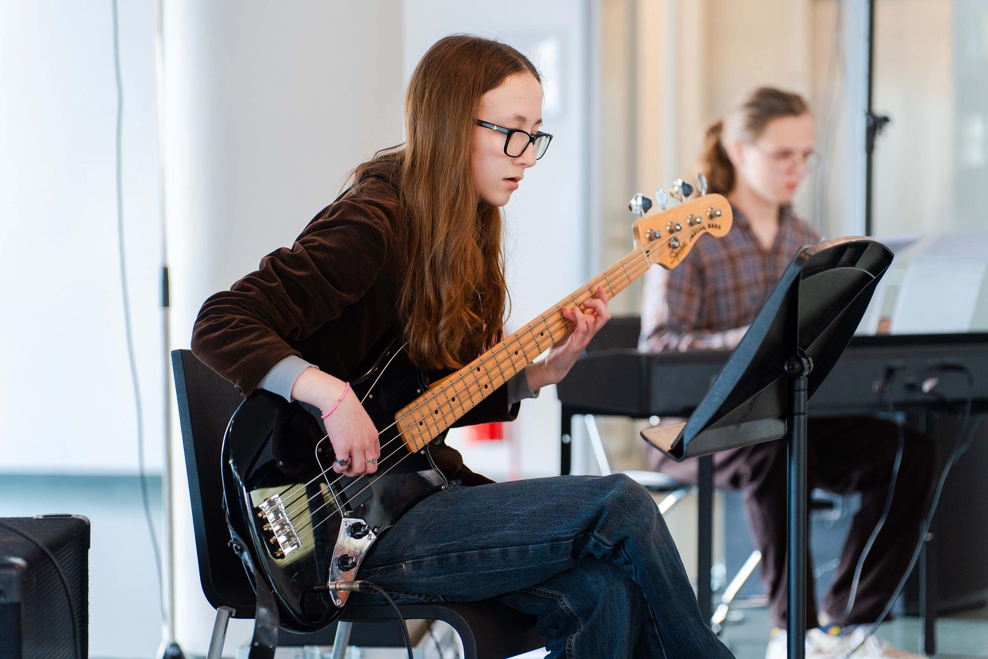 Jonge muzikant speelt elektrische basgitaar tijdens een optreden of repetitie, terwijl op de achtergrond een pianist achter een keyboard zit.
