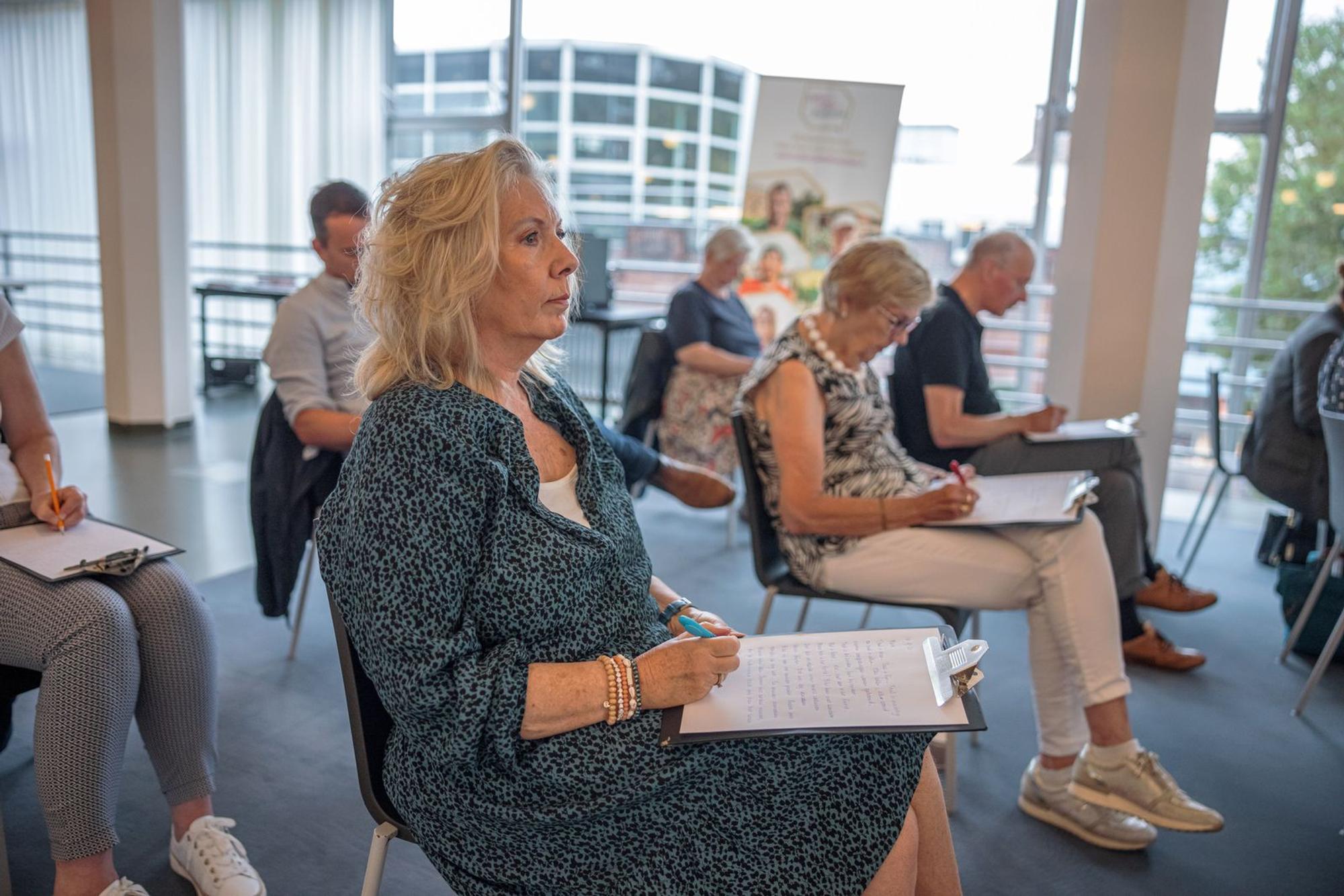 Een aantal volwassen mensen die in een rijtjesopstelling naast elkaar op een stoel zitten allen met een clipboard op hun schoot en pen in hun hand. 