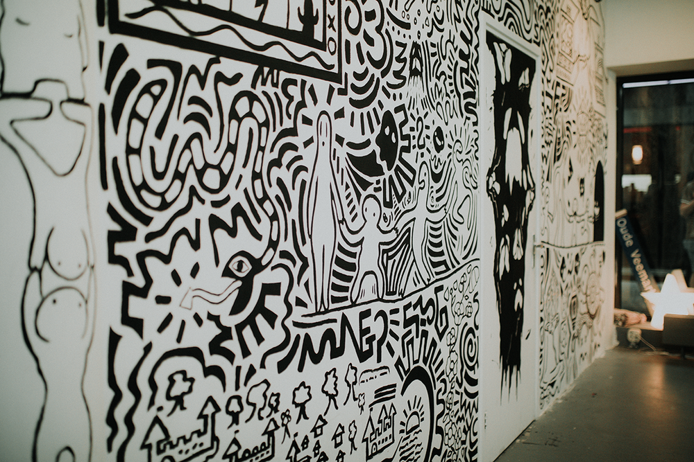 Detail van een zwart-witte muurschildering in Keith Haring-stijl, vol met abstracte figuren, patronen en symbolen op een lange muur in een binnenruimte.