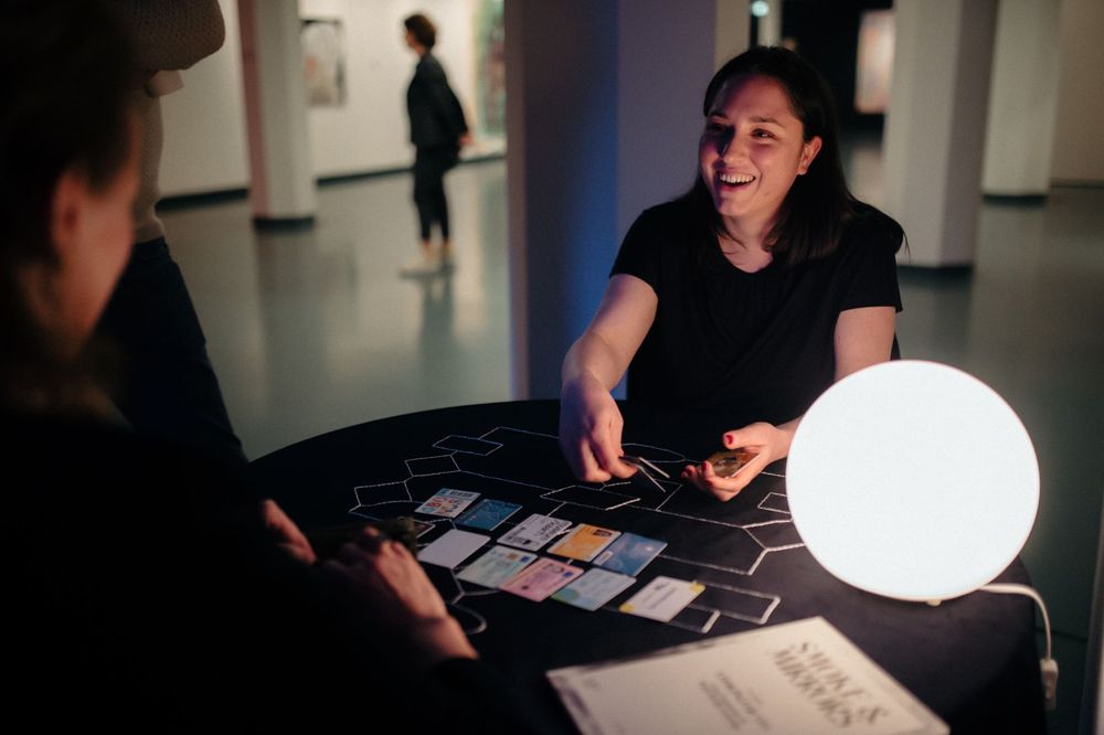 Vrouw staat aan een statafel met verschillende pasjes in haar handen. Op de tafel liggen pasjes en een bolvormige lamp. Aan de andere kant van de tafel staan twee bezoekers.