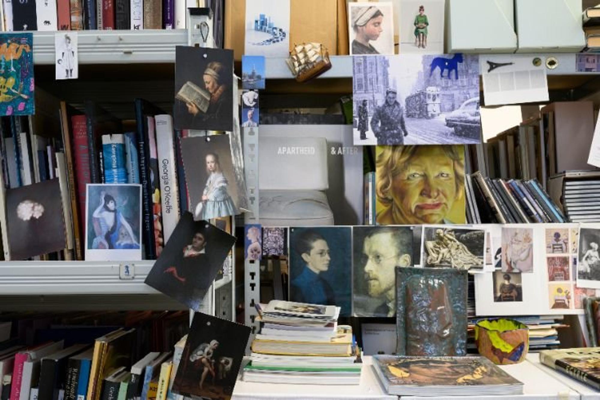 Boekenkasten in het atelier van kunstenaar Ina van Zyl, gevuld met boeken en omringd door losse afbeeldingen, foto’s en schilderijen.