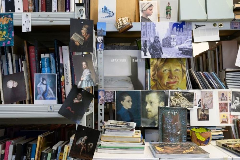 Boekenkasten in het atelier van kunstenaar Ina van Zyl, gevuld met boeken en omringd door losse afbeeldingen, foto’s en schilderijen.