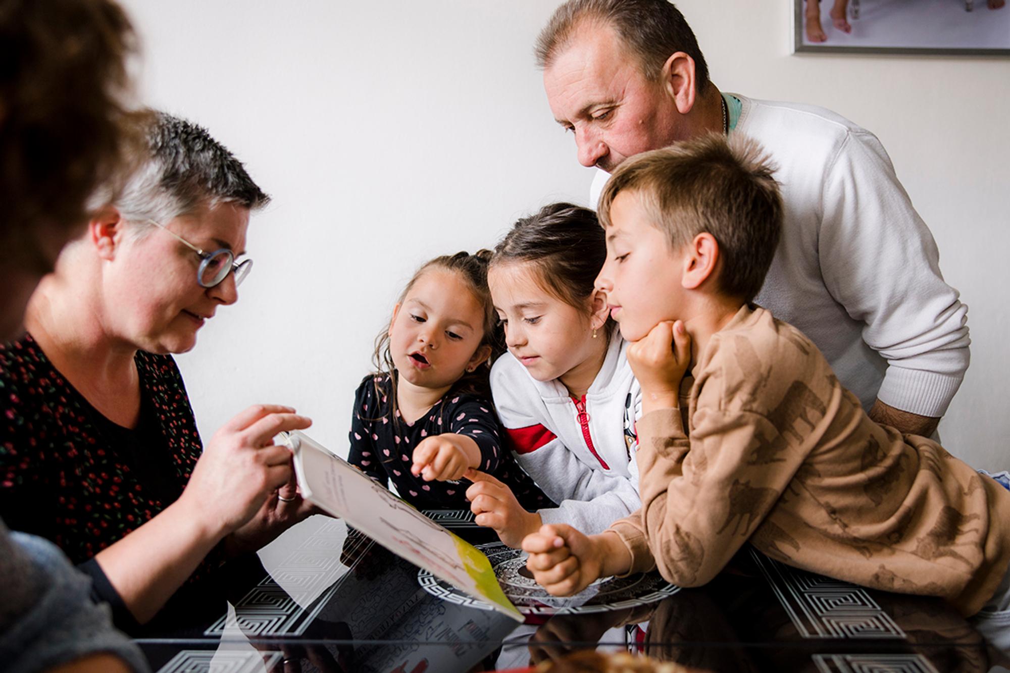 Vrijwilliger laat leesboek zien aan drie kinderen met een ouder op de achtergrond. Kinderen wijzen naar boek. 