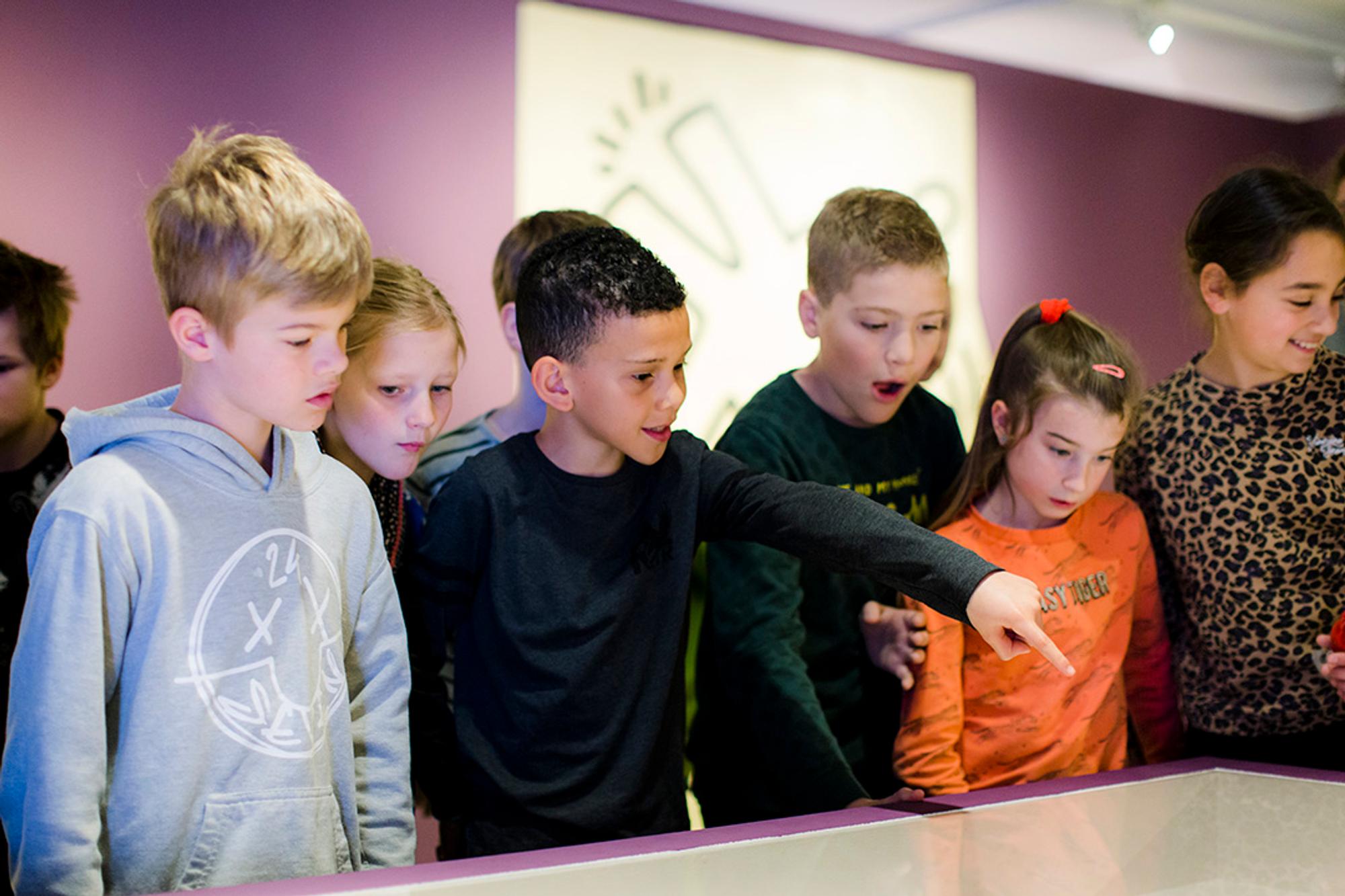 Jonge kinderen staan aandachtig voor een vitrine in de tentoonstelling ‘Keith Haring Grace House Mural’. Een van de kinderen wijst nieuwsgierig naar een object in de vitrine.

