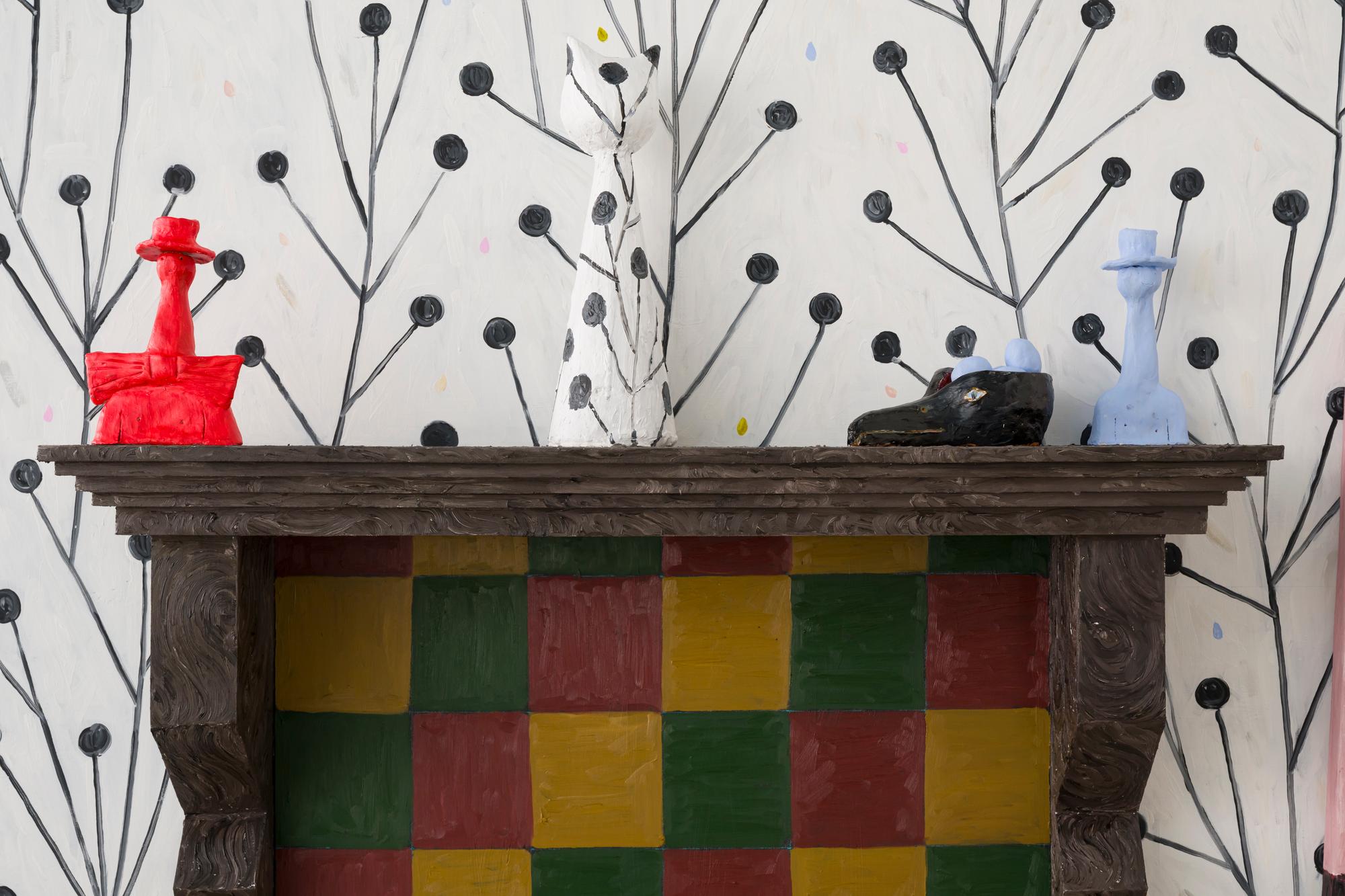 Kunstinstallatie van een schouw met rood-groen-geel geruit binnenwerk en sculpturen op de rand, waaronder een rode vaas, een wit katbeeld en een blauwe flesvorm. De muur erachter is beschilderd met een zwart takjespatroon op een witte achtergrond.