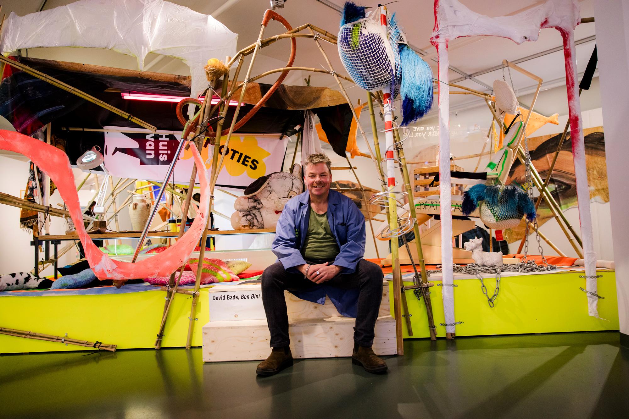 David Bade zit glimlachend voor een kleurrijke, speelse installatie van uiteenlopende materialen zoals hout, textiel en kunststof, opgebouwd als een chaotische constructie in een tentoonstellingsruimte.