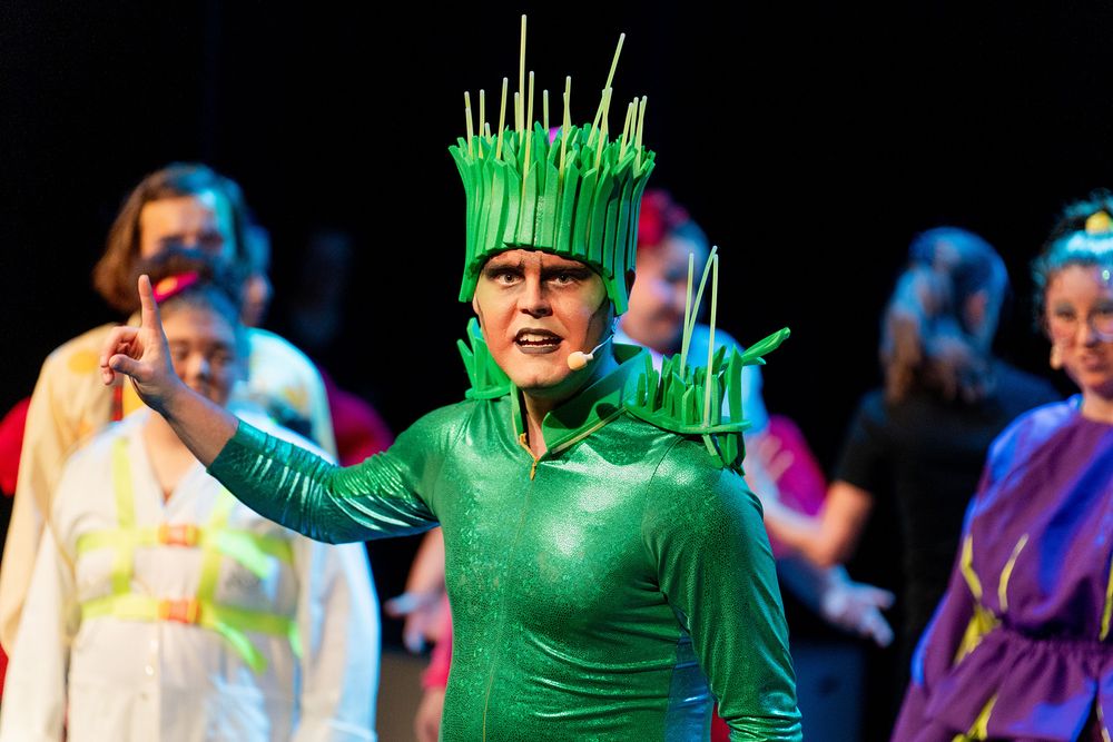 Performer in een groen kostuum met grasachtige details staat op een podium en gebaart met een opgestoken vinger, terwijl andere performers op de achtergrond staan.