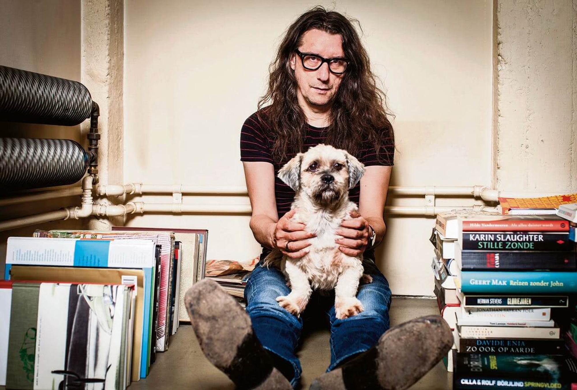 Herman Brusselmans die op de grond zit en een hondje op schoot heeft. Er staan wat boeken op de grond naast hem. 