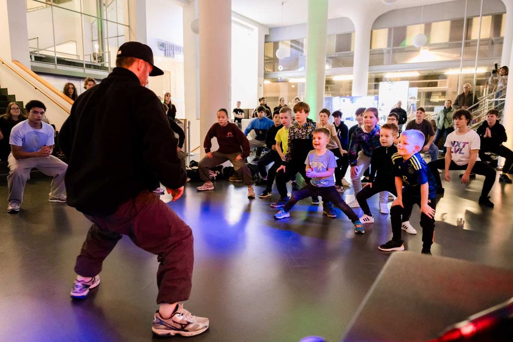 In de hal van SCHUNCK staat een man op de voorgrond, een beetje gebukt door zijn knieën, voor een groep kinderen die hem nadoen.