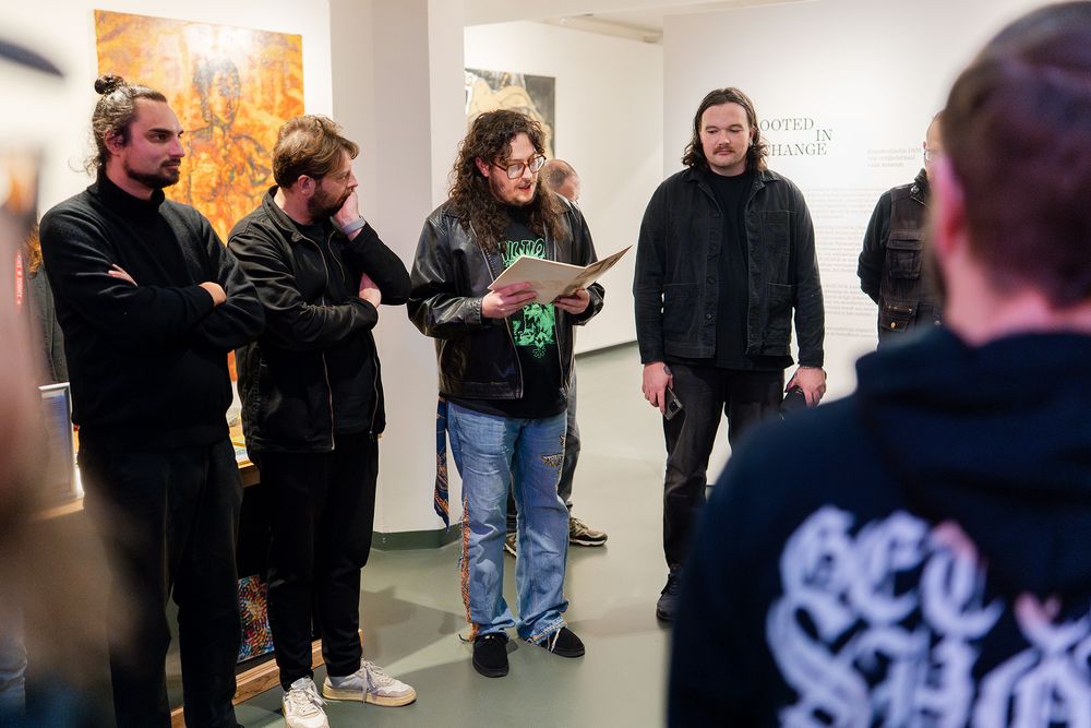 Een groep mensen staat in een galerie; in het midden leest een man met lang haar en een leren jas een tekst voor vanaf een vel papier, terwijl anderen in donkere kleding aandachtig luisteren. Op de achtergrond hangen kunstwerken en is de tekst “Rooted in Change” zichtbaar op de muur.