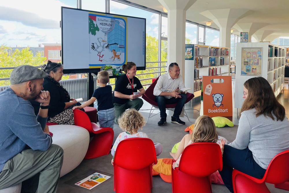 De wethouder leest voor uit Schatten van Heerlen tijdens een BoekStart-bijeenkomst in de bibliotheek. Kinderen en begeleiders zitten in een kring, terwijl op een groot scherm de boekomslag van Schatten van Heerlen wordt getoond.