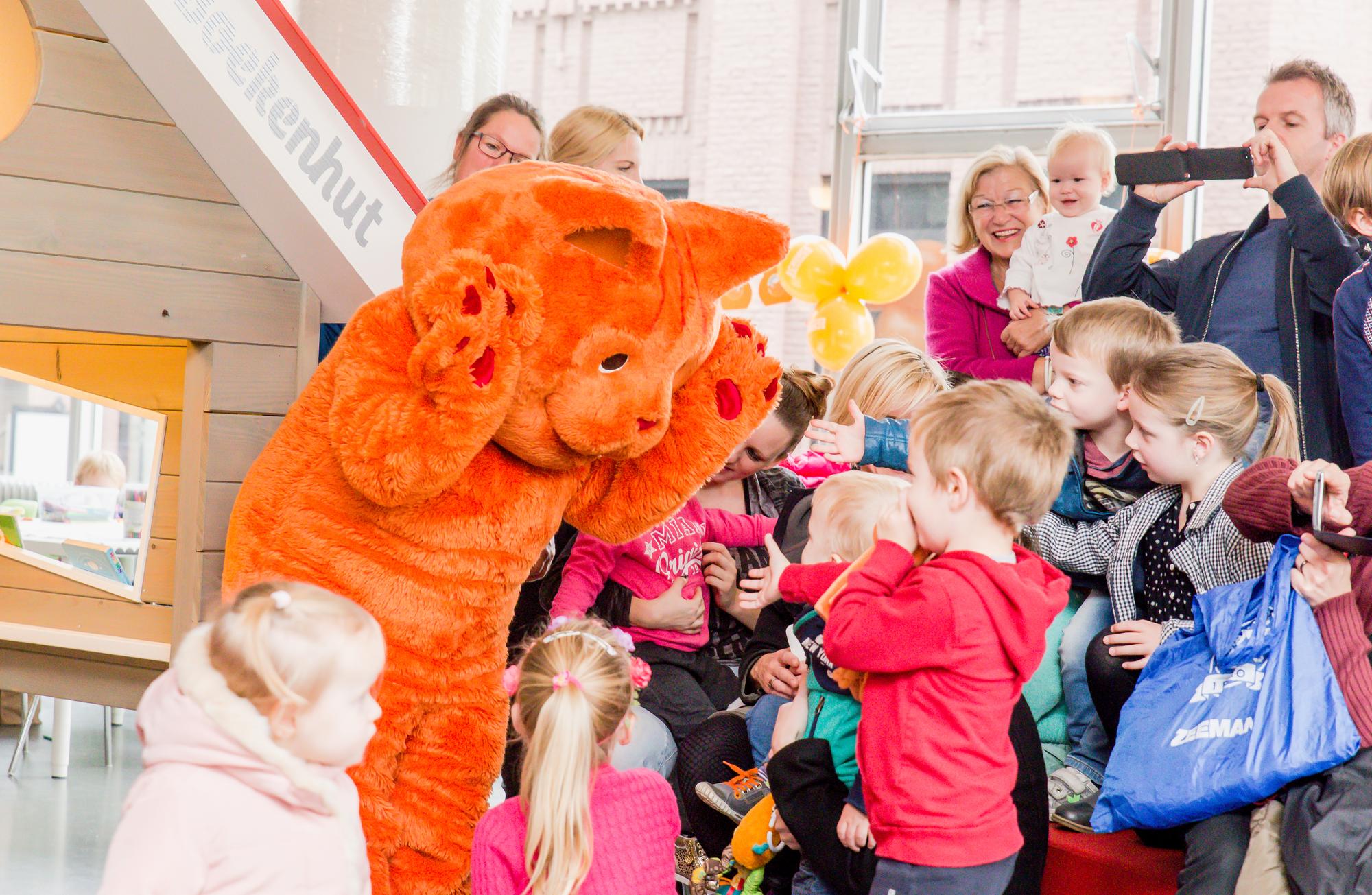 Een persoon in een groot oranje kattenpak begroet een groep enthousiaste kinderen. Op de achtergrond kijken ouders lachend toe en maken foto’s van het moment.