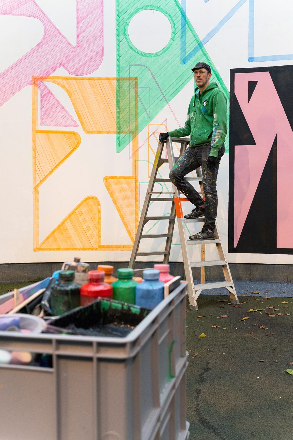 Kunstenaar Jeroen Erosie staat op een ladder voor een grote muurschildering met kleurrijke, geometrische vormen; op de voorgrond staat een bak met verfpotten.