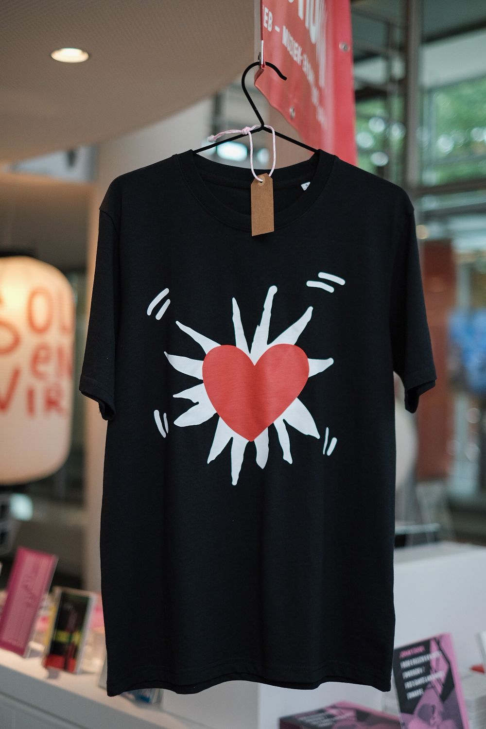 Zwart T-shirt met een ontwerp geïnspireerd op Keith Haring: een rode hartvorm in het midden, omgeven door witte stralen en bewegingslijntjes, hangend aan een hanger in een moderne winkelruimte.
