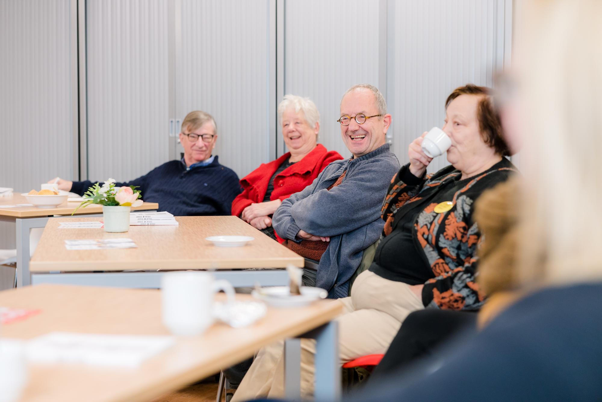 Bezoekers van Uur van de Buurt drinken lachend koffie terwijl ze aan tafels zitten. 