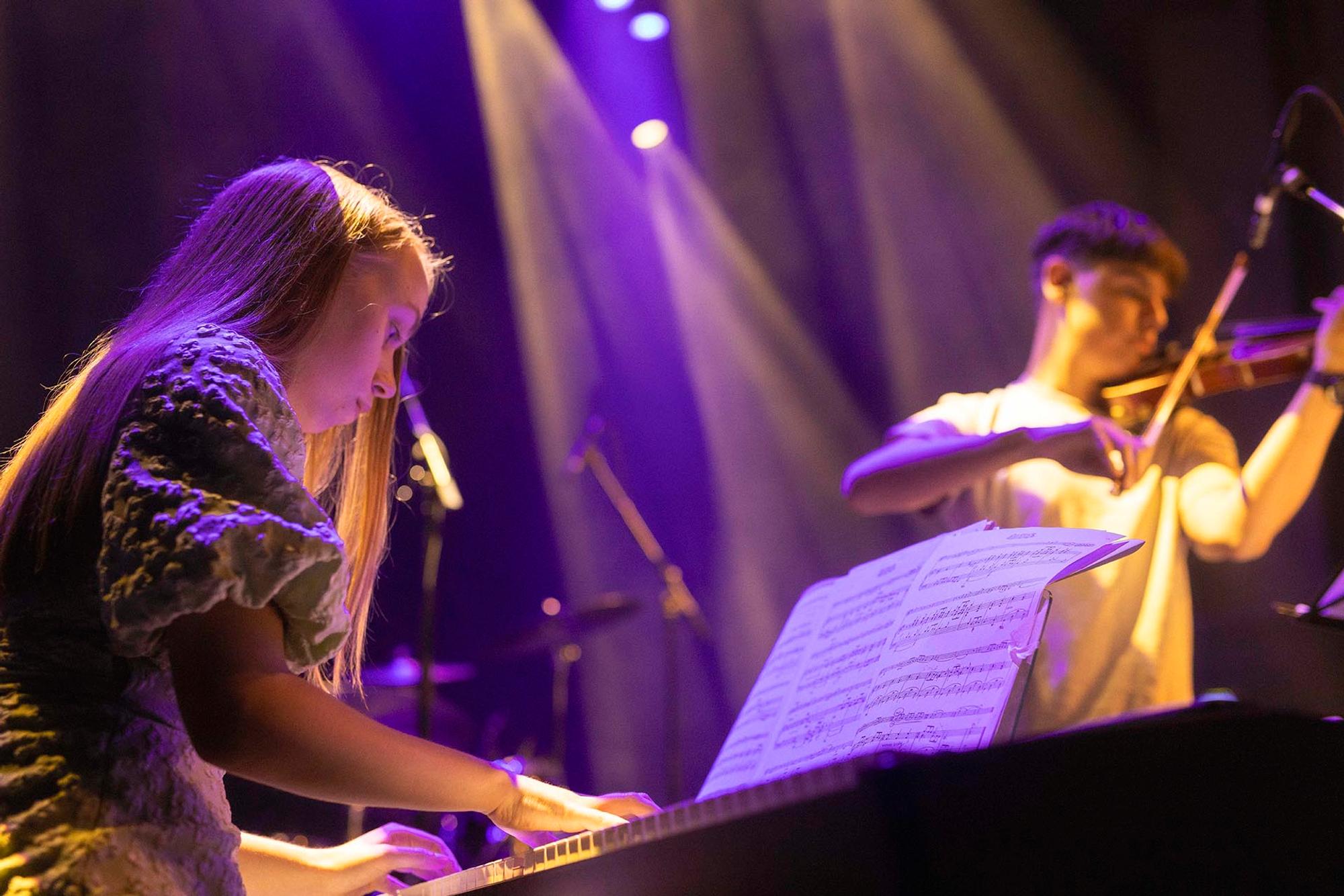 Twee jonge muzikanten op een sfeervol verlicht podium: een meisje speelt piano terwijl een jongen op de achtergrond viool speelt, met een muziekstandaard en paarse spotlichten op de achtergrond.

