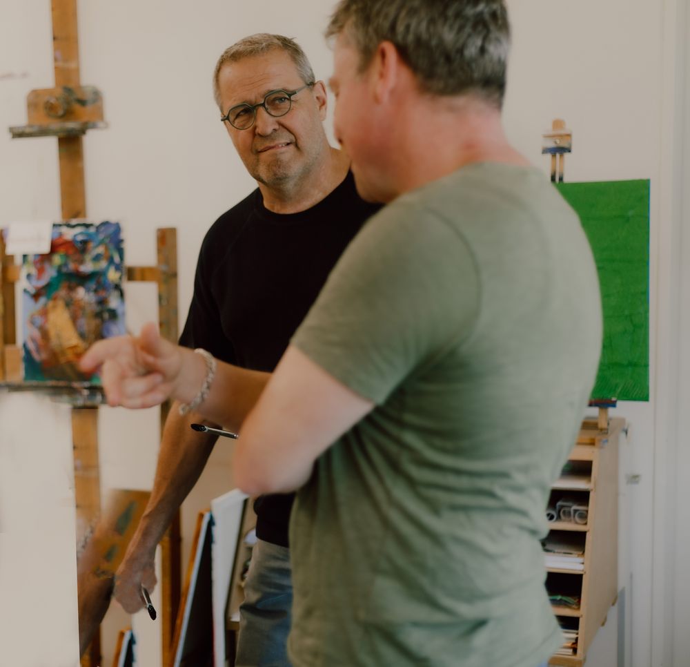 Man praat met kunstenaar David Bade, achter hem staat een ezel met een schilderij.