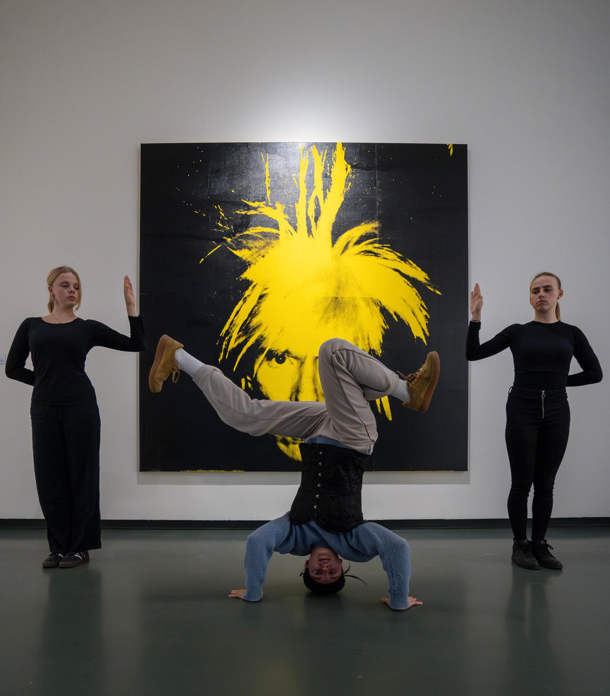 Drie studenten van Vista poseren voor een werk van Andy Warhol. De middelste student staat op zijn hoofd, tussen de twee studenten die hun hand omhoog houden.