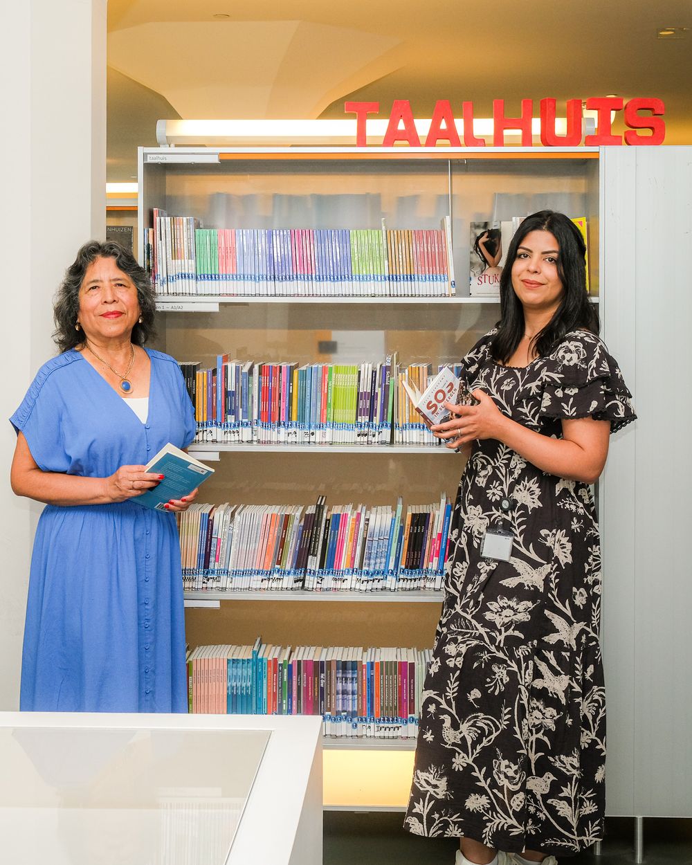 Rosario en Sara staan met boeken in hun handen voor de kast van het Taalhuis in de bibliotheek, lachend in de camera.