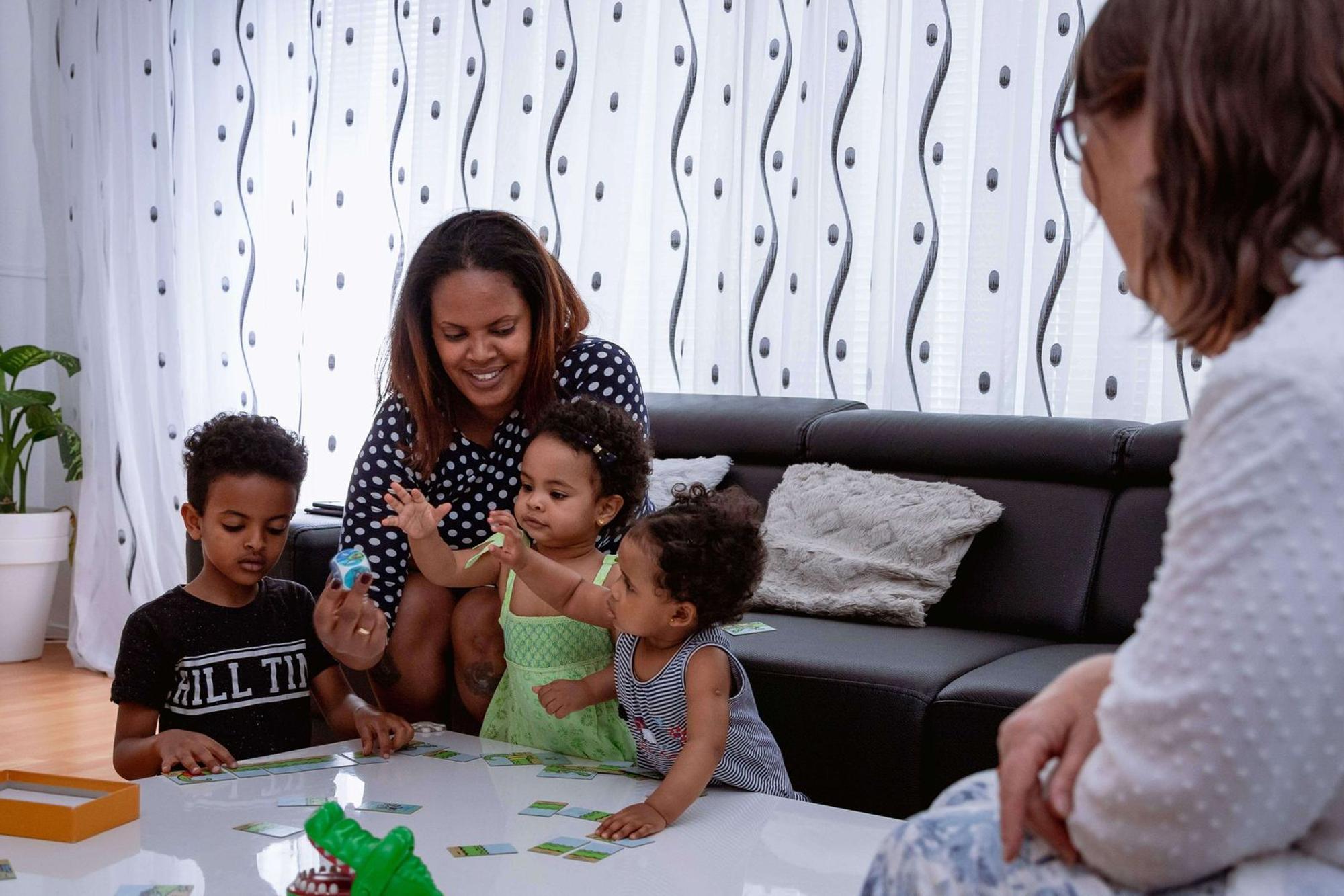 Vrouw met drie jonge kinderen speelt een spelletje aan een lage tafel. Rechts zit een vrouw en kijkt toe.