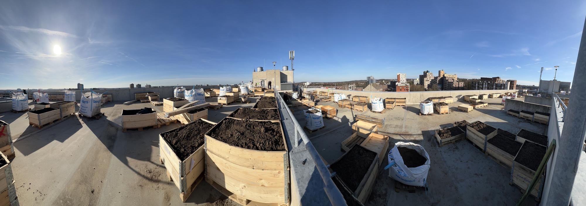 Panorama van een dakterras ingericht als moestuin, met houten bakken gevuld met aarde en grote zakken potgrond; op de achtergrond is een stad met flats en gebouwen te zien onder een heldere blauwe lucht.