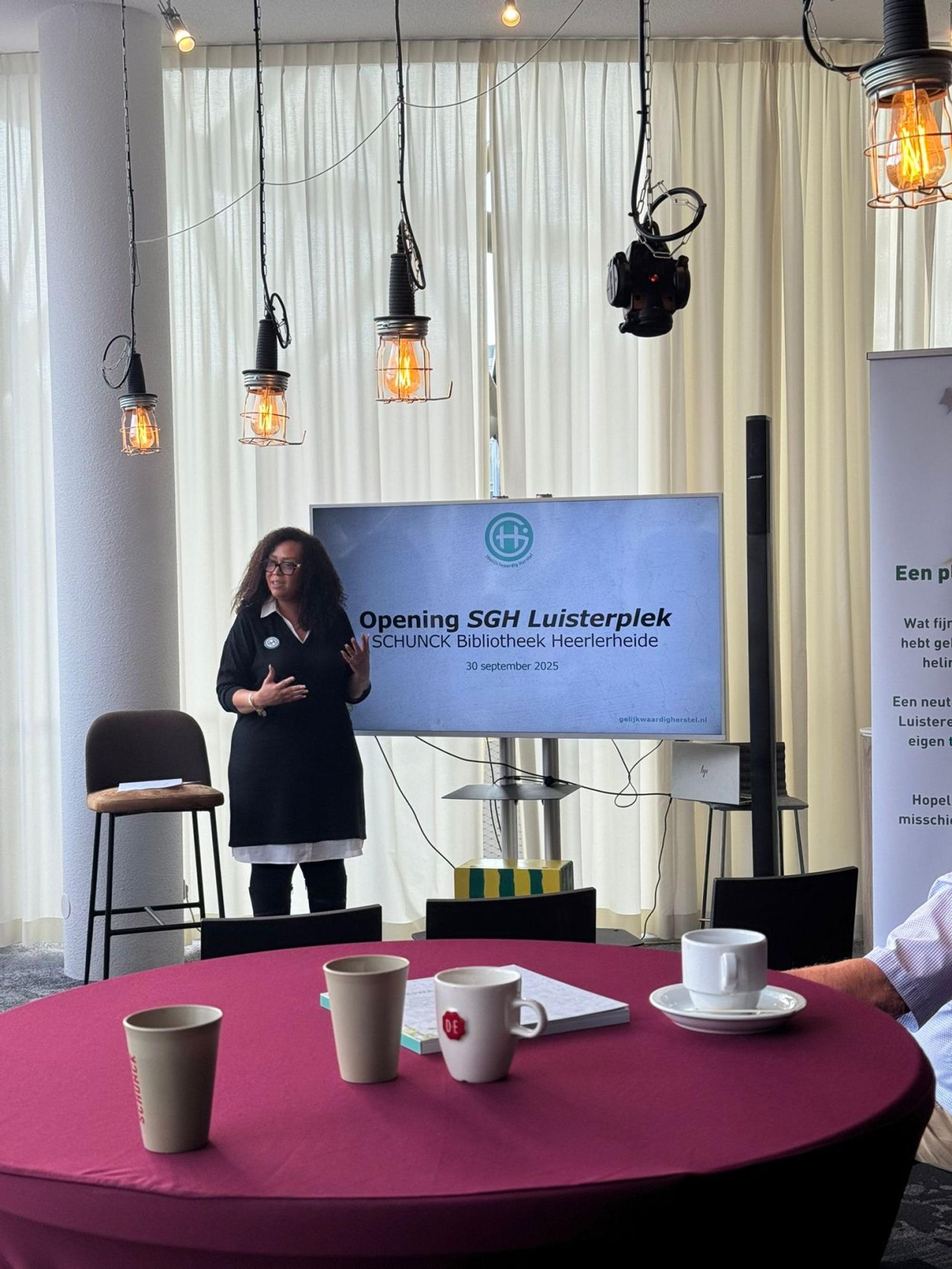 Een vrouw spreekt voor een scherm waarop staat ‘Opening SGH Luisterplek – SCHUNCK Bibliotheek Heerlerheide, 30 september 2025’. Op de voorgrond staan tafels met koffiekopjes en notitieboeken in een lichte ruimte met hanglampen.