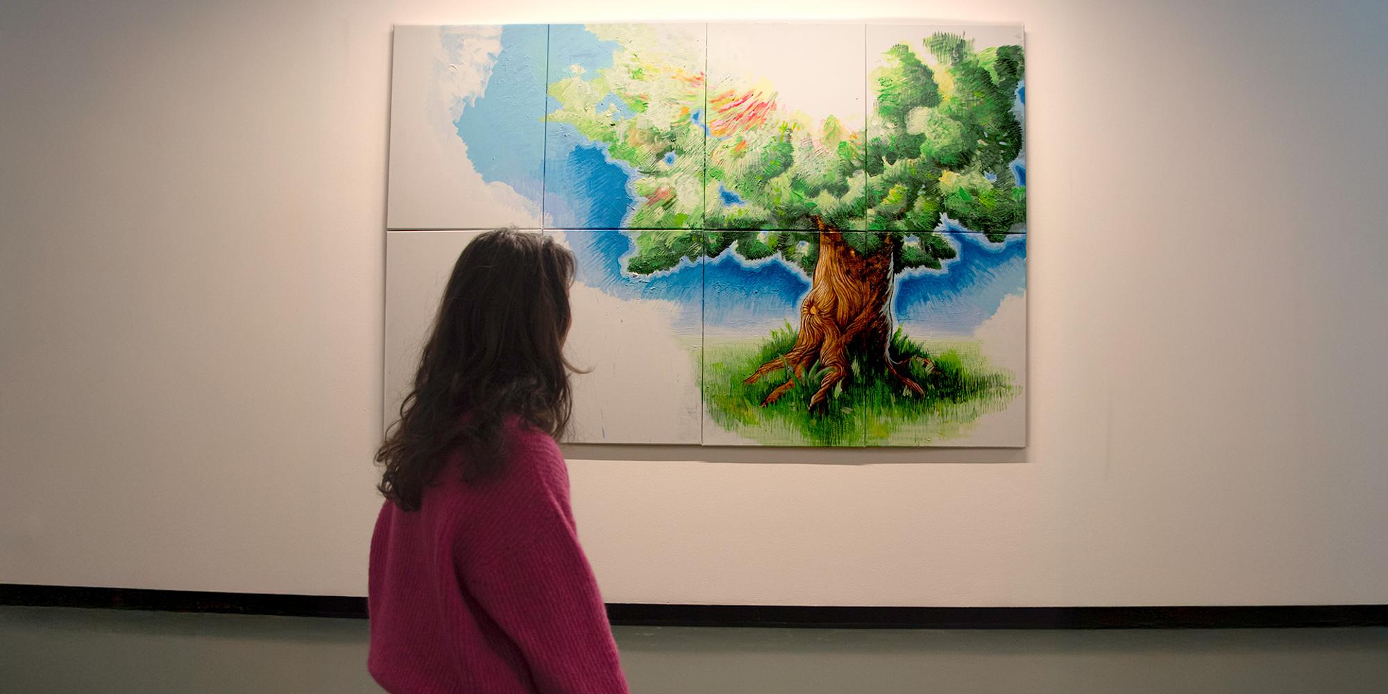 Persoon in roze trui bekijkt een kleurrijke kunstinstallatie van een boom, geschilderd op meerdere panelen tegen een witte muur.