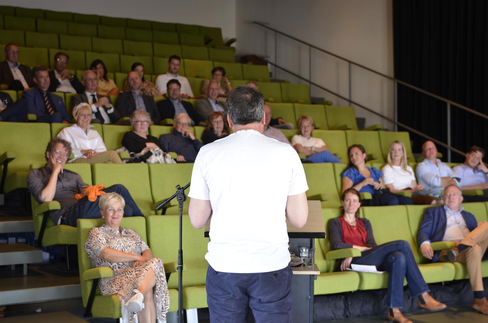 Een persoon spreekt voor een publiek in een collegezaal of auditorium. Het publiek zit op groene stoelen in rijen, en de spreker staat met de rug naar de camera. Er is een microfoonstandaard bij de spreker. De ruimte heeft een modern ontwerp met een trap zichtbaar aan de rechterkant.